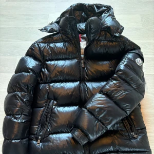 Moncler jacka  - KAN GÅ NER I PRIS VID SNABB AFFÄR! Säljer en snygg svart dunjacka från Moncler i mycket bra skick men en deffekt är att dragkedjan är lite sönder (se bild 5). Självklart äkta, skriv för äktahets bevis. Jackan är perfekt för vintern med sin pufferstil och långa ärmar. Den har en dragkedja framtill och en mysig huva. Insidan har ett coolt serietidningsmönster. Perfekt för kalla dagar! ❄️Pris kan deskuteras och skriv vid minsta lilla fundering✍️😀