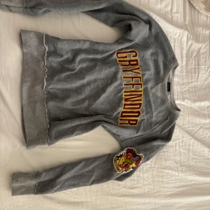 Grå Gryffindor tröja från Harry Potter - Säljer en grå tröja med Gryffindor-tryck i rött och guld. Den har långa ärmar och ett Gryffindor-märke på ärmen. Perfekt för alla Harry Potter-fans!! Aldrig använd