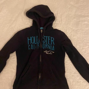 Svart hoodie från Hollister - Säljer en svart hoodie från Hollister i storlek XS. Den har en dragkedja framtill och texten 'Hollister California' i blått på bröstet. Perfekt för en avslappnad stil och mysiga dagar. 🖤