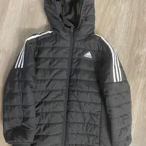 Säljer en svart pufferjacka från Adidas i mycket bra skick. Jackan har en dragkedja framtill och en huva för extra skydd mot väder. Den är tillverkad i 100% återvunnen polyester och har de klassiska Adidas-ränderna på ärmarna. Perfekt för höst och vinter! Jackan är endast använd en gång. Storleken är M i barnstorlek och är perfekt för dig som är 10-12 år.