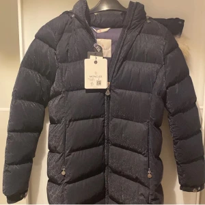 Svart dunjacka från Moncler - Snygg svart pufferjacka från Moncler med pälsdetalj på huvan. Jackan har en dragkedja framtill och långa ärmar. Perfekt för kalla vinterdagar. 