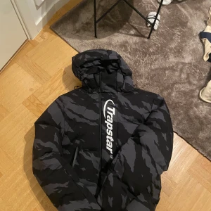 Svart och grå pufferjacka från Trapstar - Säljer en svart och grå pufferjacka från Trapstar med ett camouflage-mönster. Jackan har en stor huva och dragkedja framtill. Perfekt för kyliga dagar. Jackan är size s kan möjligtiv passa i mellan m och s var för liten för mig tyvärr så därför den säljes