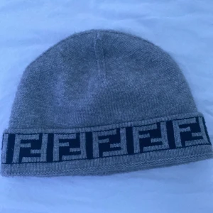 Grå mössa från Fendi - Snygg grå mössa från Fendi med ett ikoniskt mönster i svart runt kanten. Perfekt för att hålla sig varm med stil under kyliga dagar. KVITTO FINNS