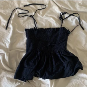 Svart axelbandslös topp - Snygg svart axelbandslös topp med knytband. Perfekt för en somrig look. Den har en smockad överdel och en lätt volang nedtill.