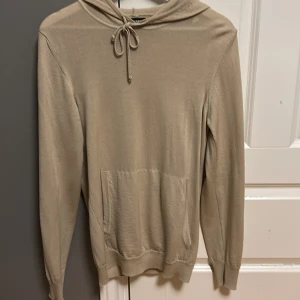 Beige hoodie från J.Lindeberg - Säljer en stilren beige hoodie från J.Lindeberg i merinoull. Tröjan har en justerbar huva med snörning och ribbade muddar vid ärmslut och nederkant. Perfekt för en avslappnad och bekväm stil. Ända defekt på tröjan är att fickan har gått av lite på kanten (se bild)