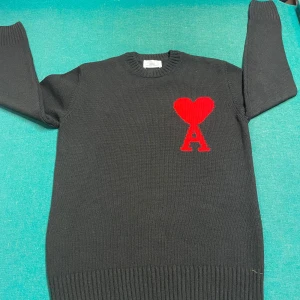 Svart tröja från Ami Alexandre Mattiussi - Snygg svart tröja från Ami Alexandre Mattiussi med ett rött hjärta och bokstaven A på bröstet. Tröjan är stickad och har långa ärmar med ribbade muddar. Perfekt för en stilren look.