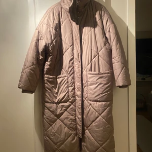 Beige quiltad kappa - Säljer en stilren beige quiltad kappa med lång ärm och dragkedja. Stor i storleken. Perfekt för kyliga dagar med sin långa passform och praktiska fickor framtill. 