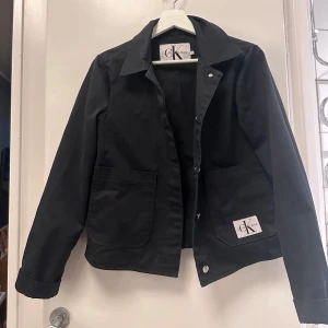 Svart jacka från Calvin Klein Jeans - Snygg svart jacka från Calvin Klein Jeans med klassisk krage och knappar framtill. Jackan har två stora fickor och en stilren design som passar perfekt för olika tillfällen.