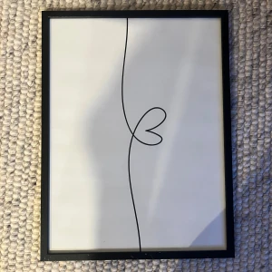 Tavla med hjärtmotiv - En stilren tavla med en enkel linjeteckning av ett hjärta i svart mot vit bakgrund. Ramen är svart och rektangulär, vilket ger en modern och minimalistisk känsla. Perfekt för att ge en subtil konstnärlig touch till ditt hem.