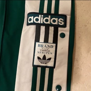Gröna Adidas byxor med knappar - Snygga gröna Adidas byxor med vita och svarta detaljer. Byxorna har knappar längs sidorna och de klassiska tre ränderna. Perfekta för en sportig look.