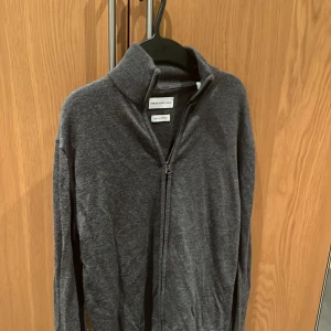 Grå merinoulltröja från These Glory Days - Säljer en stilren grå tröja i merinoull från These Glory Days. Tröjan har en dragkedja framtill och en hög krage, perfekt för kyliga dagar. Den är långärmad och har en mjuk och bekväm passform.