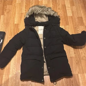 Svart dunjacka från Ralph Lauren - Snygg svart dunjacka från Ralph Lauren med pälskantad huva. Jackan har långa ärmar och stängs med dragkedja och knappar. Perfekt för kalla vinterdagar. 