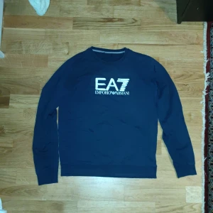 Mörkblå tröja från Emporio Armani - Säljer en stilren mörkblå tröja från Emporio Armani med EA7-logga på bröstet. Tröjan har långa ärmar och rund halsringning. Perfekt för en avslappnad och trendig look. Har defekt vid armen Obs! Kolla bild 3. Stl S men skulle passa 160-165