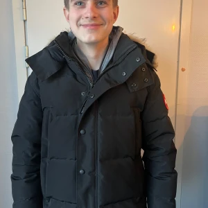 Svart dunjacka med pälsdetalj - Säljer en svart dunjacka från Canada Goose med pälsdetalj på huvan. Jackan har en klassisk design med lång ärm och dragkedja framtill. Perfekt för kalla vinterdagar.