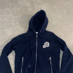 Mörkblå hoodie med dragkedja - Snygg mörkblå hoodie med dragkedja och huva. Den har ett broderat märke på bröstet och praktiska fickor med dragkedja. Perfekt för en avslappnad stil.