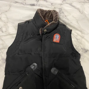 Svart dunväst från Parajumpers - Snygg svart dunväst från Parajumpers med en mysig fuskpälsfodrad krage. Västen har dragkedja och knappar framtill samt praktiska fickor med dragkedjor. Perfekt för kyliga dagar.
