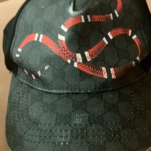 Svart keps - Snygg svart keps med ett iögonfallande rött ormmönster. Kepsen har ett diskret mönster i bakgrunden och är perfekt för att ge din outfit en cool touch.