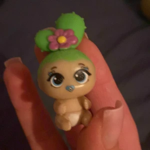 En liten docka med kaktus hår  - En liten figur med grönt hår och en rosa blomma på huvudet. Figuren har stora ögon och en napp i munnen. Den bär en vit blöja och har en söt, tecknad stil.