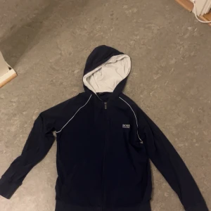 Mörkblå hoodie från Hugo Boss - Säljer en stilren mörkblå hoodie från Hugo Boss med dragkedja och vit insida i huvan. Perfekt för en avslappnad look. Den har långa ärmar och en klassisk passform.
