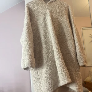 Beige teddyhoodie från Kappahl - Mysig beige teddyhoodie från Kappahl med huva och dekorativa bollar. Perfekt för kyliga dagar med sin fluffiga och varma design. Hoodien har en avslappnad passform och är idealisk för höst och vinter.