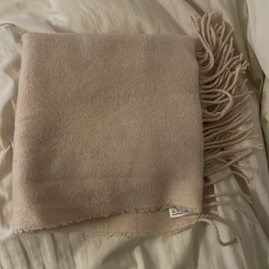 Beige halsduk från H&M - Säljer en stilren beige halsduk från H&M med fransar i ändarna. Perfekt för att hålla dig varm under kyliga dagar. Den är mjuk och bekväm att bära.