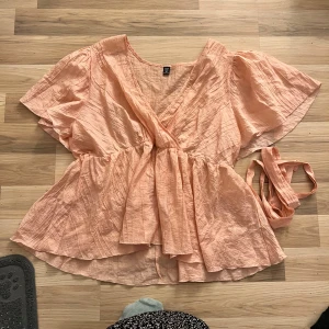Persikofärgad omlottblus från Shein - Säljer en söt persikofärgad omlottblus från Shein i storlek 4XL. Blusen har korta ärmar och ett knytband i midjan för en fin passform. Perfekt för vår och sommar!
