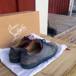 Grå/blå sneakers från Christian Louboutin - Säljer ett par grå/blå sneakers från Christian Louboutin i okej skick. Skorna har den ikoniska röda sulan och blå snörning. Perfekta för en stilren look med en touch av lyx. Kommer med originalkartong.