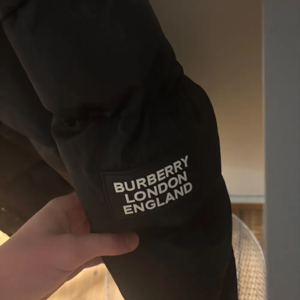 Säljer en snygg svart dunjacka från Burberry med deras klassiska rutiga mönster på insidan av huvan. Jackan har en dragkedja och knappar framtill, perfekt för kalla vinterdagar. Den är i mycket bra skick och har en stilren design med Burberry-logga på ärmen. Passar både till vardags och fest!. Jackor.