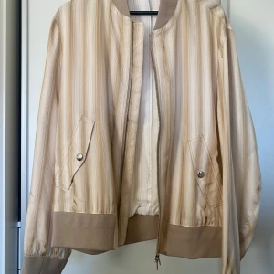 Beige randig bomberjacka från Tiger of Sweden - Säljer en snygg beige bomberjacka från Tiger of Sweden. Den har ett stilrent randigt mönster och praktiska fickor med knappar. Perfekt för vår och höst! Jackan har en dragkedja framtill och ribbade muddar vid ärmslut och nederkant. Passar både till vardags och fest.