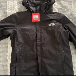 Svart regnjacka från The North Face - Säljer en helt ny svart regnjacka från The North Face. Den har en praktisk huva och dragkedja framtill. Perfekt för regniga dagar och håller dig torr och bekväm. Jackan är i nytt skick och har en stilren design med The North Face-loggan på bröstet.