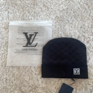 Svart mössa från Louis Vuitton - Snygg och stilren svart mössa från Louis Vuitton i nytt skick. Den har en diskret logga framtill och är perfekt för både höst och vinter. Mössan är gjord i ett mjukt material som håller dig varm och bekväm. Ett måste för den modeintresserade!