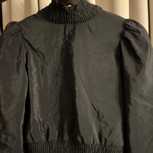 Svart blus med hög krage och knyt - Svart blus med hög krage och puffiga ärmar. Knytning och öppen rygg i midjan. Använd 1 gång, bra skick. Pris kan diskuteras🩷