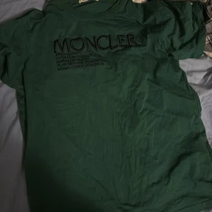 Grön t-shirt från Moncler - Säljer en snygg grön t-shirt från Moncler i bra skick. Den har korta ärmar och ett tryck med Moncler-loggan på framsidan. Perfekt för en avslappnad stil eller vardagsbruk. Materialet känns mjukt och bekvämt, perfekt för vår och sommar! Sänks vid snabb affär