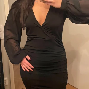 Svart omlottklänning - Snygg svart omlottklänning med v-ringning och långa, puffiga ärmar. Klänningen har en tight passform som framhäver figuren och är perfekt för festliga tillfällen. Materialet är mjukt och stretchigt, vilket gör den bekväm att bära hela kvällen.