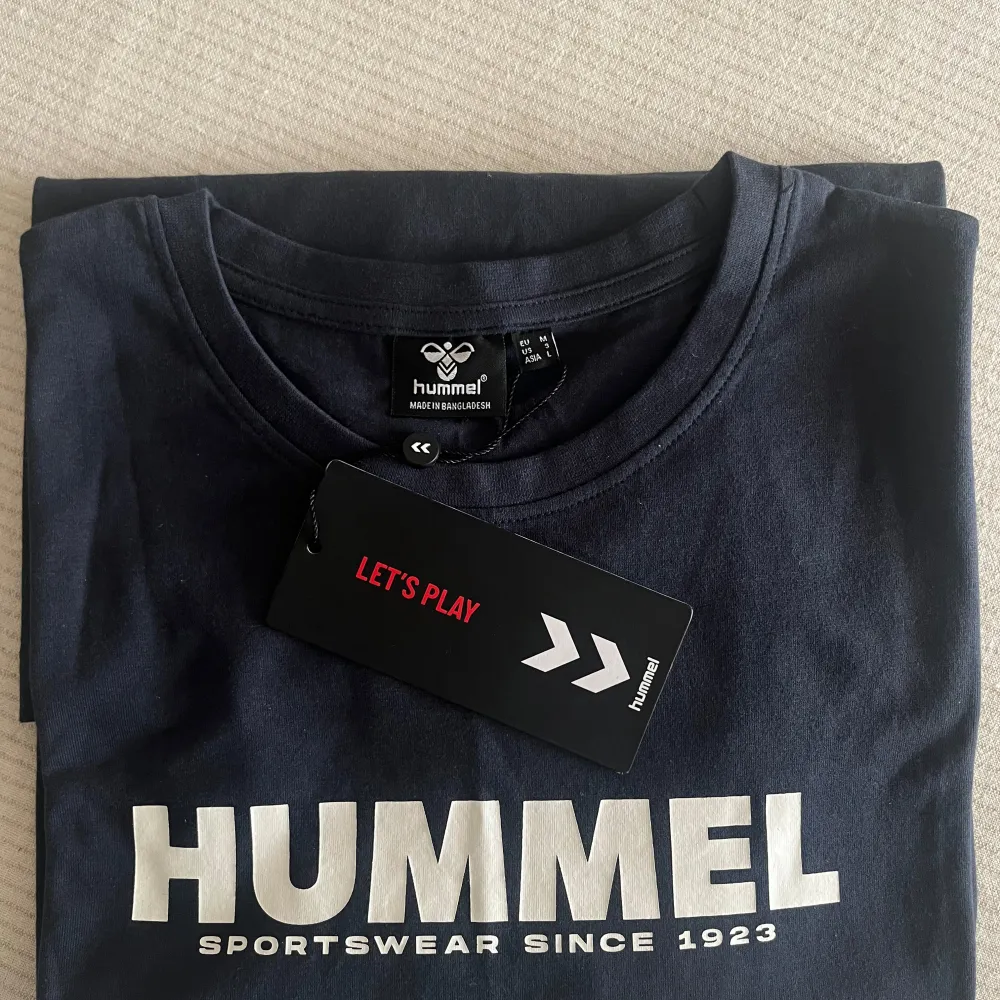 Säljer en mörkblå t-shirt från Hummel i mjukt, stickat jerseytyg. Den har en vit logga framtill med texten 'Hummel Sportswear Since 1923'. Perfekt för både träning och vardag. Unisex design för en bekväm passform.. T-shirts.