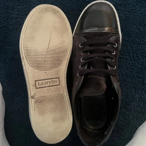 Gråa/bruna  sneakers från Lanvin - Säljer ett par sneakers från Lanvin i bra skick. Skorna har en snygg glansig tå och klassisk snörning. Perfekta för både vardag och fest. Sulan är vit med Lanvin-loggan. Passar till många olika outfits! Pris kan diskuteras vid snabb affär, köparen får både med ett extra snöre och ett mocka kitt som har använts en gång, mocka kittet kan användas många gånger, hör av er vid minsta fråga✋