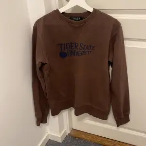 Storlek M men passar S.Säljer en brun sweatshirt från Tiger of Sweden med trycket 'Tiger State University' i blått på framsidan. Tröjan har långa ärmar och en klassisk rund halsringning. Perfekt för en avslappnad stil eller en dag på campus. Skön och stilren!