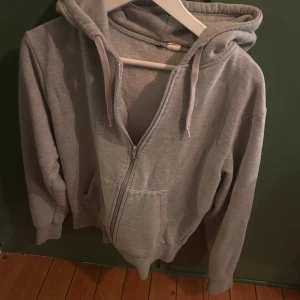Grå hoodie med dragkedja - Säljer en grå hoodie i bra skick. Den har en dragkedja framtill och en mysig huva med snören. Perfekt för kyliga dagar eller som ett extra lager. Passar till det mesta och är superbekväm!