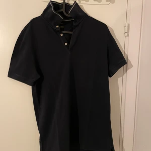 Svart pikétröja från Massimo Dutti - Snygg svart pikétröja från Massimo Dutti i bra skick. Den har en klassisk krage med knappar och korta ärmar, perfekt för både vardag och lite finare tillfällen. Materialet känns mjukt och bekvämt mot huden.