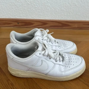 Vita Nike Air Force 1 sneakers - Säljer ett par klassiska vita Nike Air Force sneakers i bra skick. De har en stilren design med snörning och perforerade detaljer på ovansidan för extra ventilation. Perfekta för både vardag och fest! 