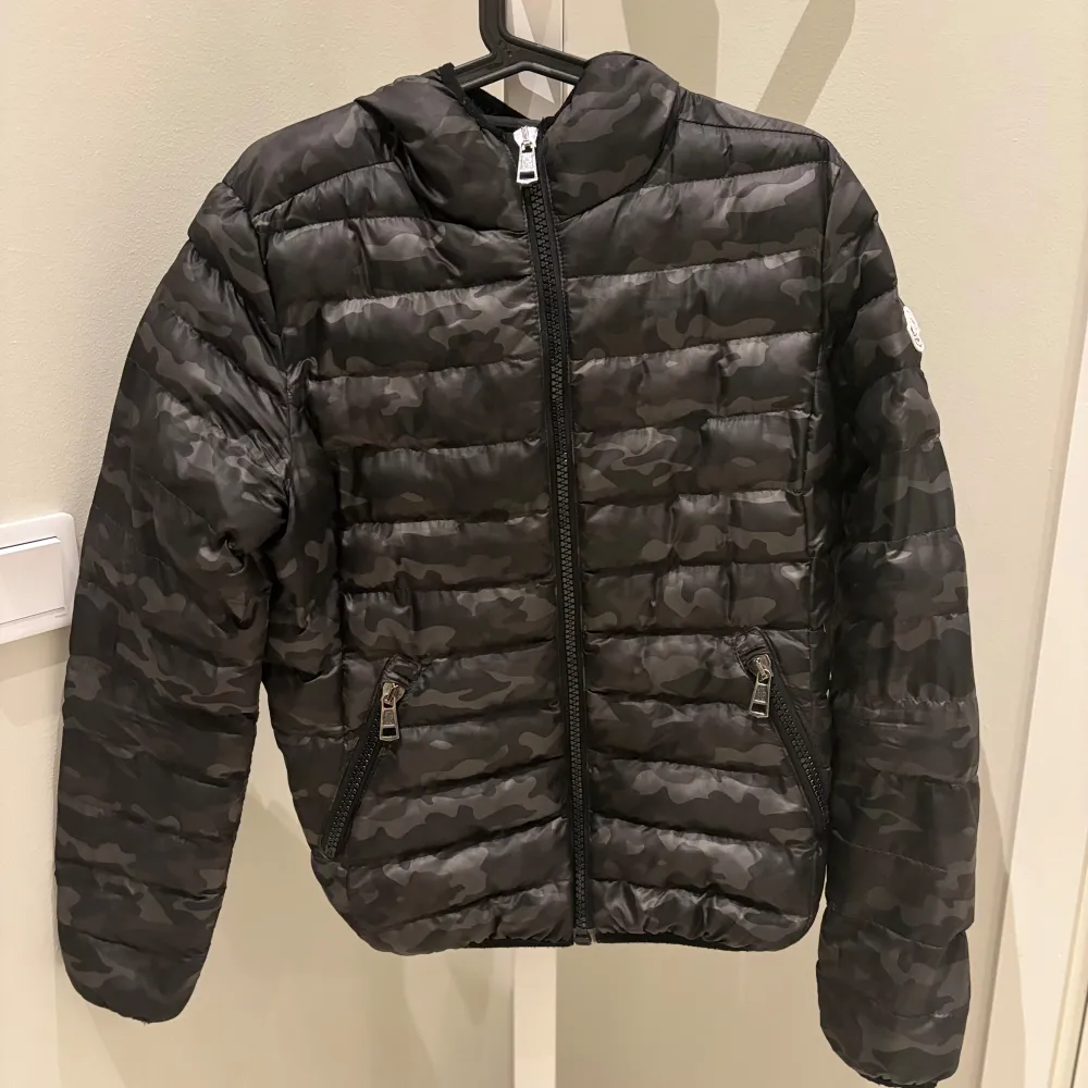 Säljer en snygg svart camo dunjacka från Moncler. Jackan har en cool camouflage-look och är perfekt för höst och vinter. Den har en dragkedja framtill och två praktiska fickor med dragkedjor. Perfekt för att hålla sig varm och stilren! 🖤. Takit.