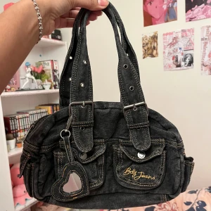 Svart axelväska från Baby Jeans - Säljer en cool svart axelväska från Baby Jeans i jeansmaterial. Väskan har flera fickor med knappar och en snygg hjärtformad nyckelring. Perfekt för vardagsbruk och ger en avslappnad stil. Den har dubbla handtag och en dragkedja för säker förvaring.