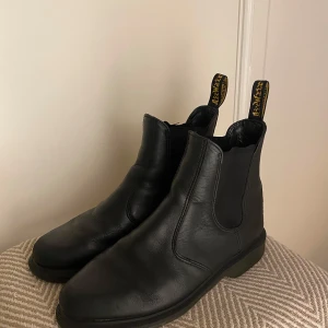 Dr. Martens Svarta Chelsea boots - Säljer ett par klassiska svarta Laura Chelsea boots från Dr. Martens i skinn. De har den ikoniska gula tagen och en bekväm, slitstark sula. De är i använt men bra skick, de har många år kvar att ge. 