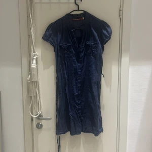 Mörkblå klänning med knappar - Säljer en mörkblå klänning med knappar framtill. Den har korta ärmar och ett knytband i midjan för en justerbar passform. Perfekt för både vardag och festliga tillfällen!