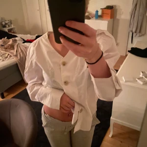 Vit blus med knappar - Säljer en stilren vit blus med knappar framtill. Den har långa ärmar och en lös passform, perfekt för både vardag och fest. Blusen är i mycket bra skick och passar till många olika outfits. Perfekt till alla tillfällen!