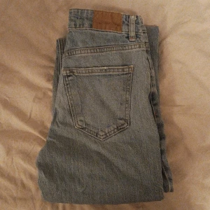 Blåa jeans från Gina Tricot  - Säljer ett par grå jeans från Perfect Jeans i storlek 34. De har en klassisk design med fem fickor och knappgylf. Jeansen är i bra skick med en liten sliten detalj på benet som ger dem en cool look. Perfekta för vardagsbruk! Perfekt passform för dom som vill framhäva kurvor