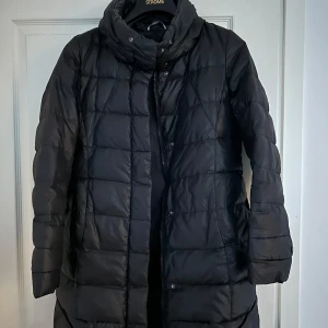 Svart dunjacka från Max Mara - Säljer en elegant svart dunjacka från Max Mara. Jackan har en quiltad design med långa ärmar och en hög krage för extra värme. Den har en dragkedja och knappar framtill samt praktiska fickor. Från kollektion år 2002, köpt second hand. Storlek XL barn, passar S dam. 