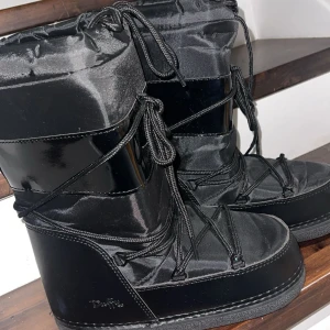 Svarta Moon Boots från Duffy - Säljer ett par svarta Moon Boots från Duffy i mycket bra skick. De är gjorda i syntetmaterial med snörning och har en robust sula perfekt för vintern. Bootsens glansiga yta ger en cool look och de är supervarma och bekväma. Perfekta för kalla vinterdagar! ❄️