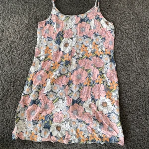 Blommig sommarklänning med knytband - Söt blommig klänning perfekt för sommardagar! Den har ett färgglatt mönster med rosa, orange och vita blommor. Klänningen är ärmlös med smala axelband som knyts, vilket ger en justerbar passform. Materialet känns lätt och luftigt, perfekt för varma dagar.