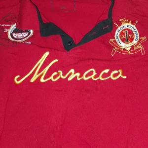 Röd pikétröja med broderi - Säljer en snygg röd pikétröja med broderad text 'Monaco' i guld på framsidan. Tröjan har en klassisk krage och knappdetalj framtill. Perfekt för en avslappnad men stilren look. Passar bra till både vardag och fest! OBS LITEN STORLEK 